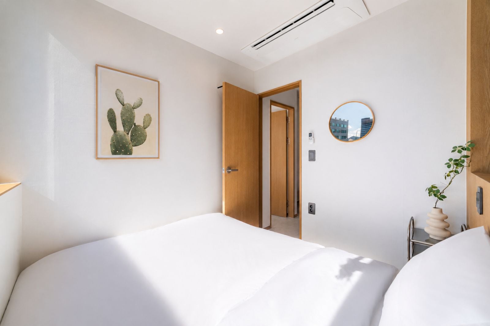 A modern hotel room with a bed, a door leading to another room, a wall-mounted mirror, and a cactus artwork.