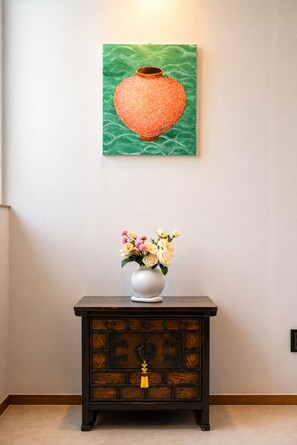 Un tableau représentant un objet en forme de cœur, accroché au mur sur un fond vert, un vase blanc rempli de fleurs posé sur une commode en bois foncé en dessous, et un interrupteur sur le mur, à droite.