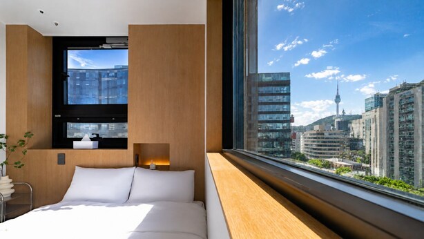 Una habitación de hotel moderna con una cama grande, una mesita de noche, un ventanal con vista a la ciudad y un panel de pared de madera.