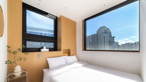 Habitación de hotel moderna con una cama grande, una ventana con vistas a la ciudad, una mesita de noche y una planta pequeña.