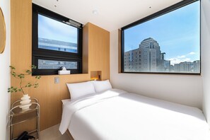 Une chambre d’hôtel moderne avec un grand lit, une fenêtre donnant sur la ville, une table de chevet et une petite plante.