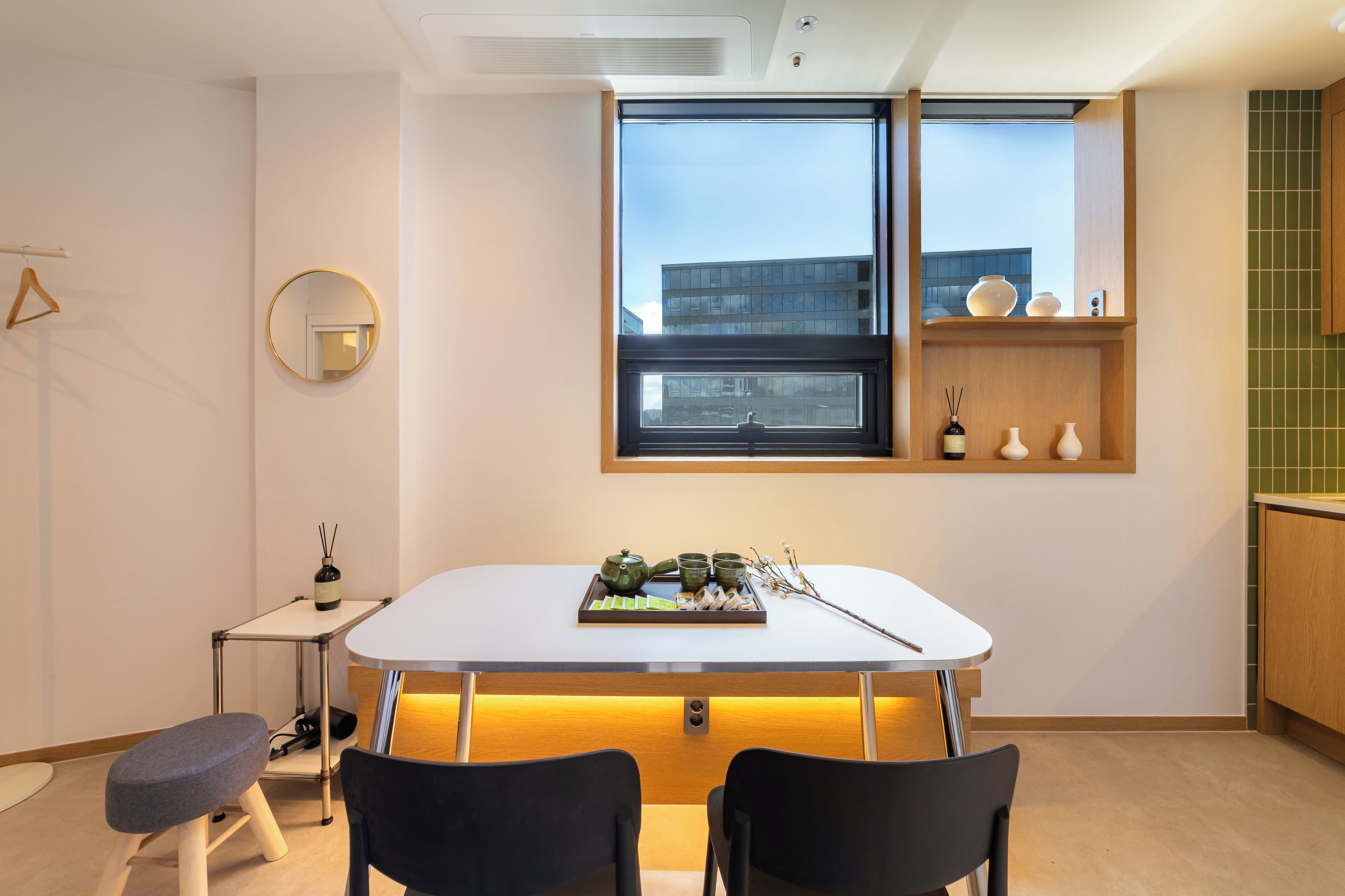 A modern kitchen with a dining table, chairs, a window with a view of buildings, and a small shelf with decorative items.