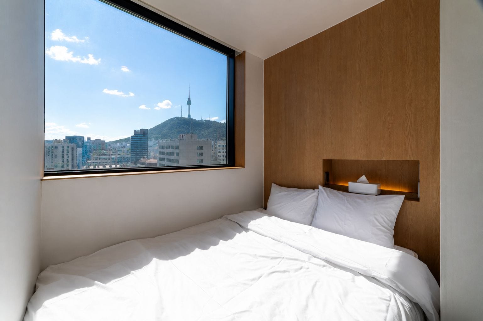 Habitación de hotel con cama, cabecera de madera, mesita de noche y ventana con vista a la ciudad.