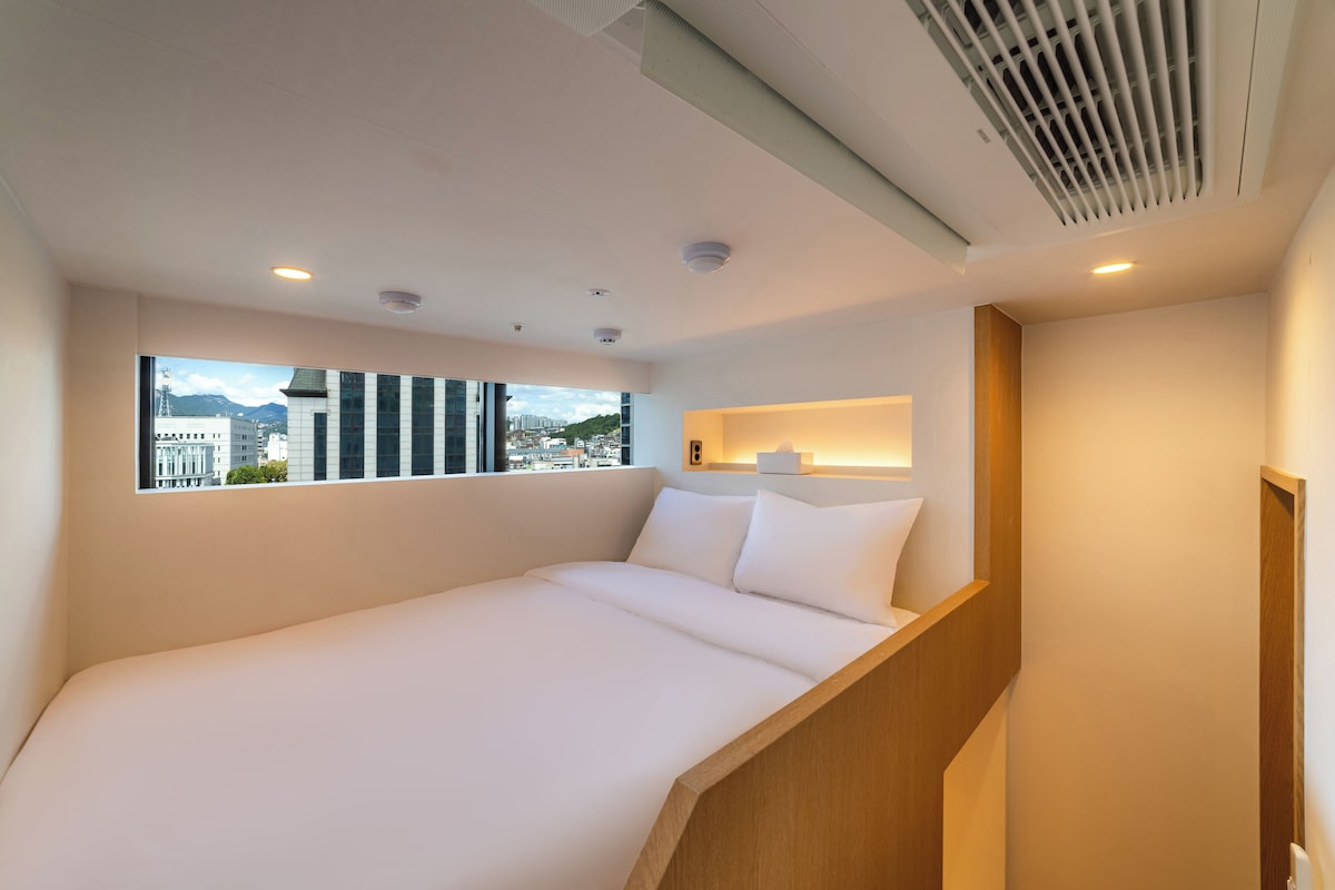 Une chambre d’hôtel avec un lit, une fenêtre donnant sur la ville, un escalier en bois et un luminaire fixé au mur.
