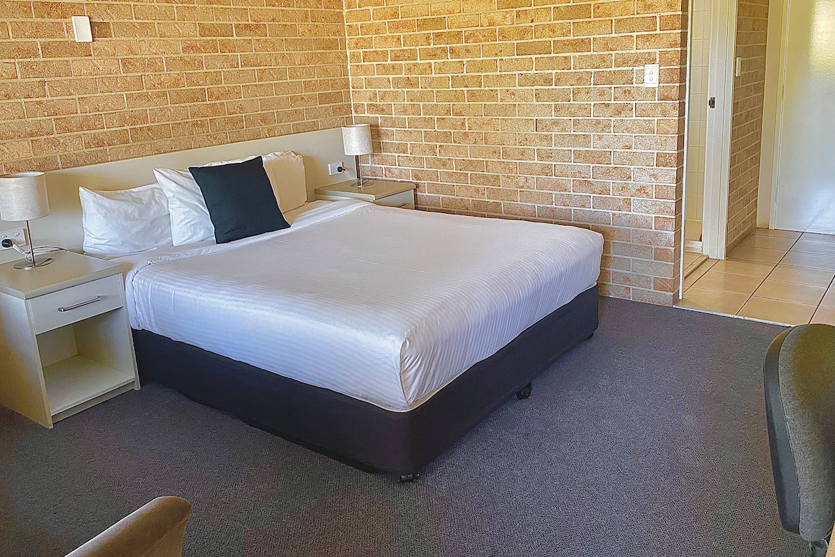 Ein Hotelzimmer mit einer Ziegelwand, einem Bett mit weißer Bettwäsche, einem Nachttisch mit einer Lampe und einer Tür, die in einen anderen Raum führt.