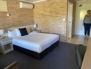Ein Hotelzimmer mit Backsteinwand, einem Bett mit weißer Bettwäsche, einem Nachttisch, einer Lampe und einer Tür zu einem weiteren Raum.