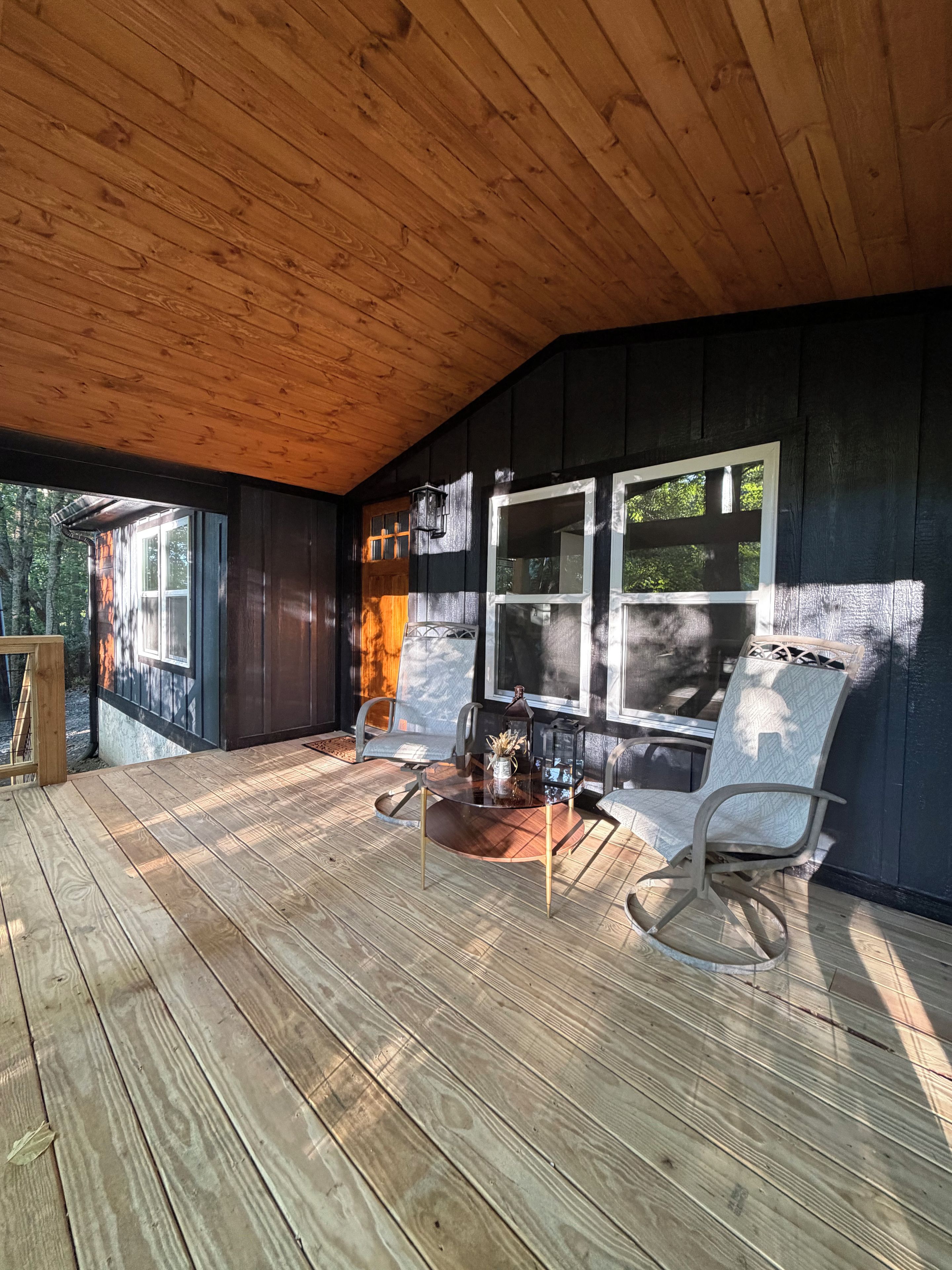 Eine überdachte Holzterrasse mit einem Glastisch und zwei Stühlen, umgeben von dunklen Holzwänden und großen Fenstern.