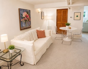 A living room with a white sofa, a glass coffee table, a round dining table with chairs, a painting on the wall, and a wooden door.