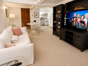A living room with a white sofa, a TV displaying a show, and a kitchen in the background.