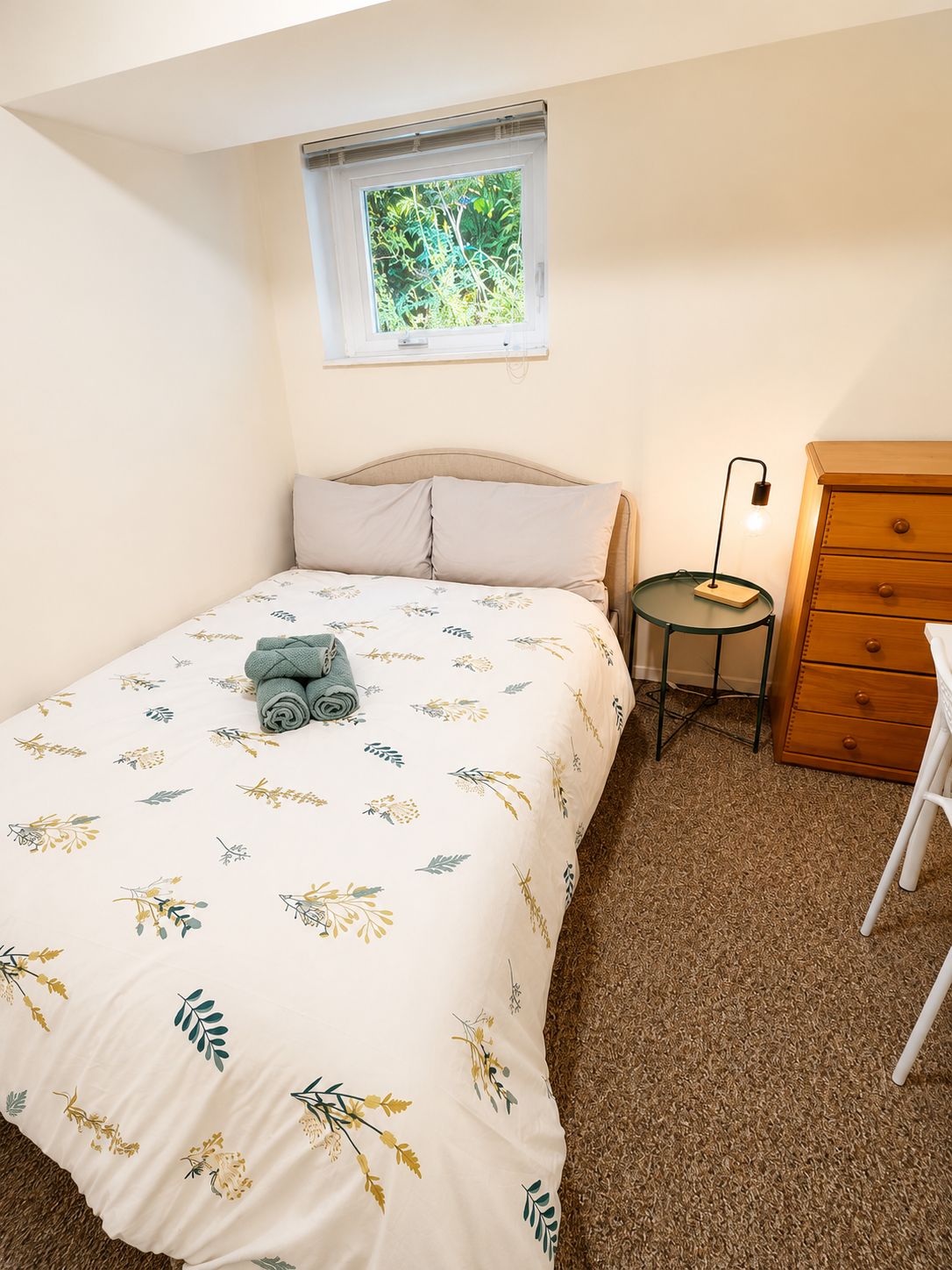 A bedroom with a bed, a small table with a lamp, a dresser, and a window with a view of greenery.