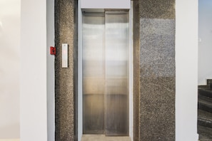 An elevator with a control panel on the left, a metallic door, and a textured wall on the right. - Super Townhouse Alwal Road (Hyderabad)