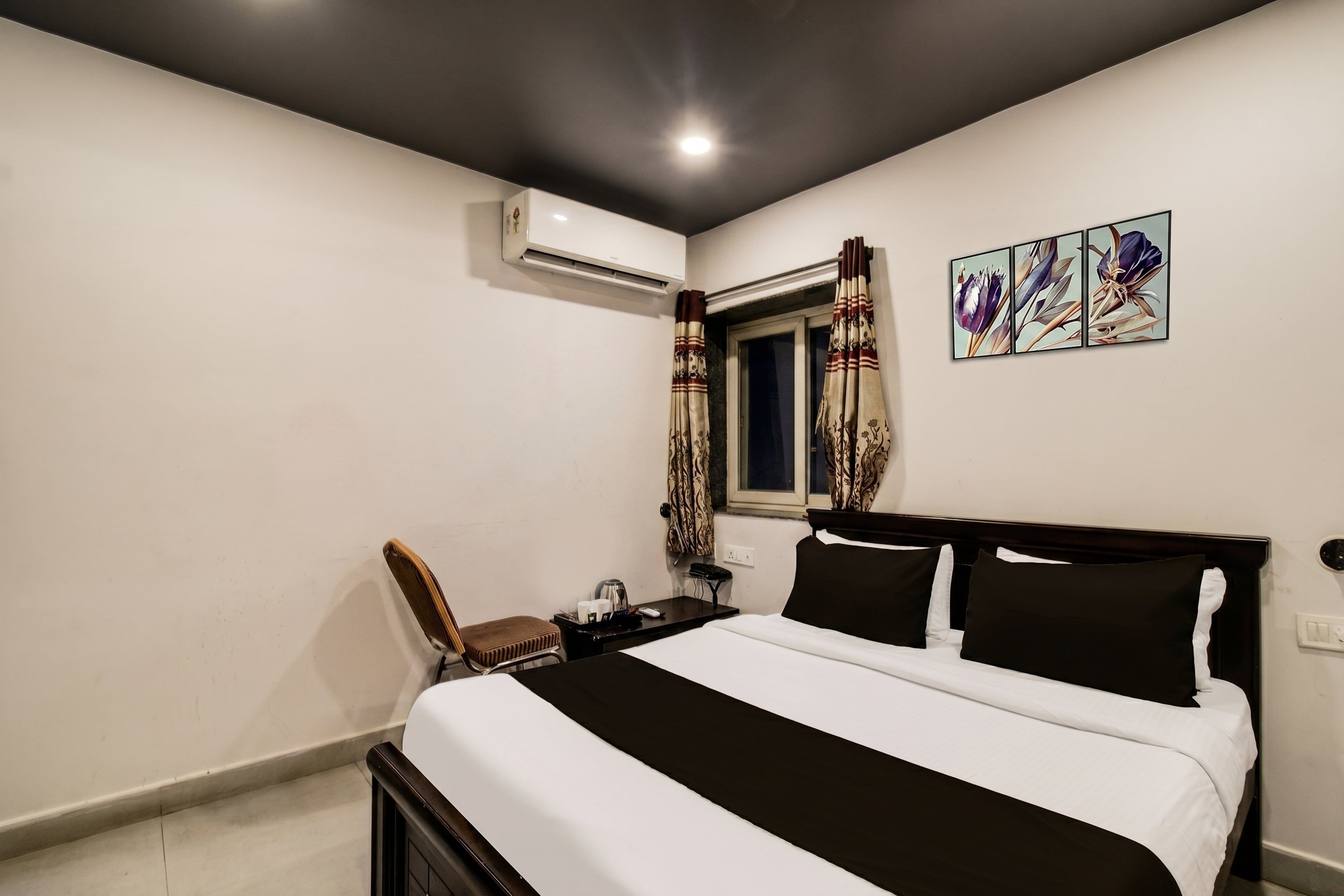 Quarto de hotel com cama, cadeira, escrivaninha, ar condicionado, janela com cortinas e uma obra de arte na parede.