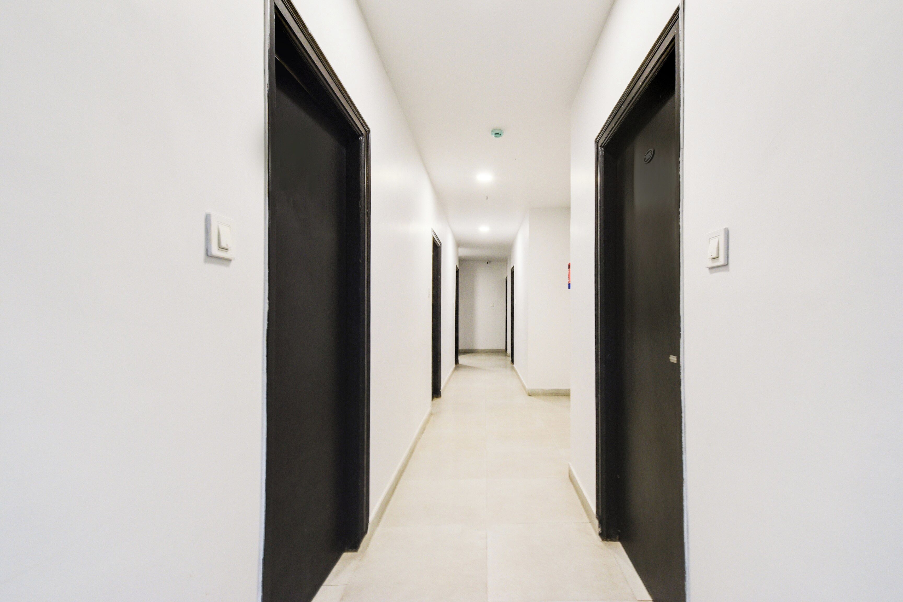 Um corredor comprido e estreito, com paredes brancas e portas pretas, que leva a um quarto no final.