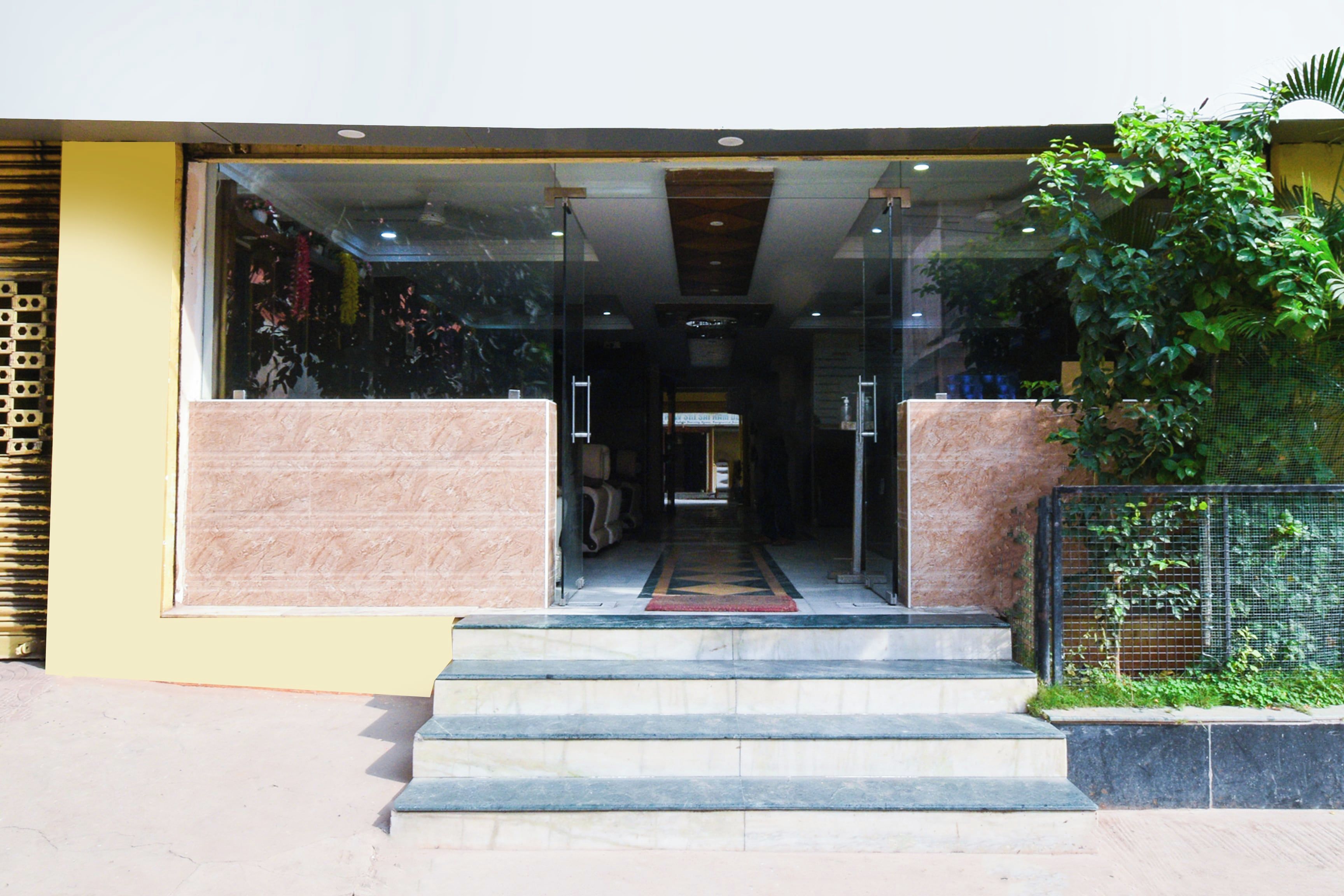 A building entrance with a glass facade, a set of stairs leading up to a door, and a small garden area to the right.