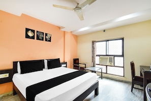 Una habitación de hotel con una cama, un ventilador de techo, una ventana con aire acondicionado y una mesita.