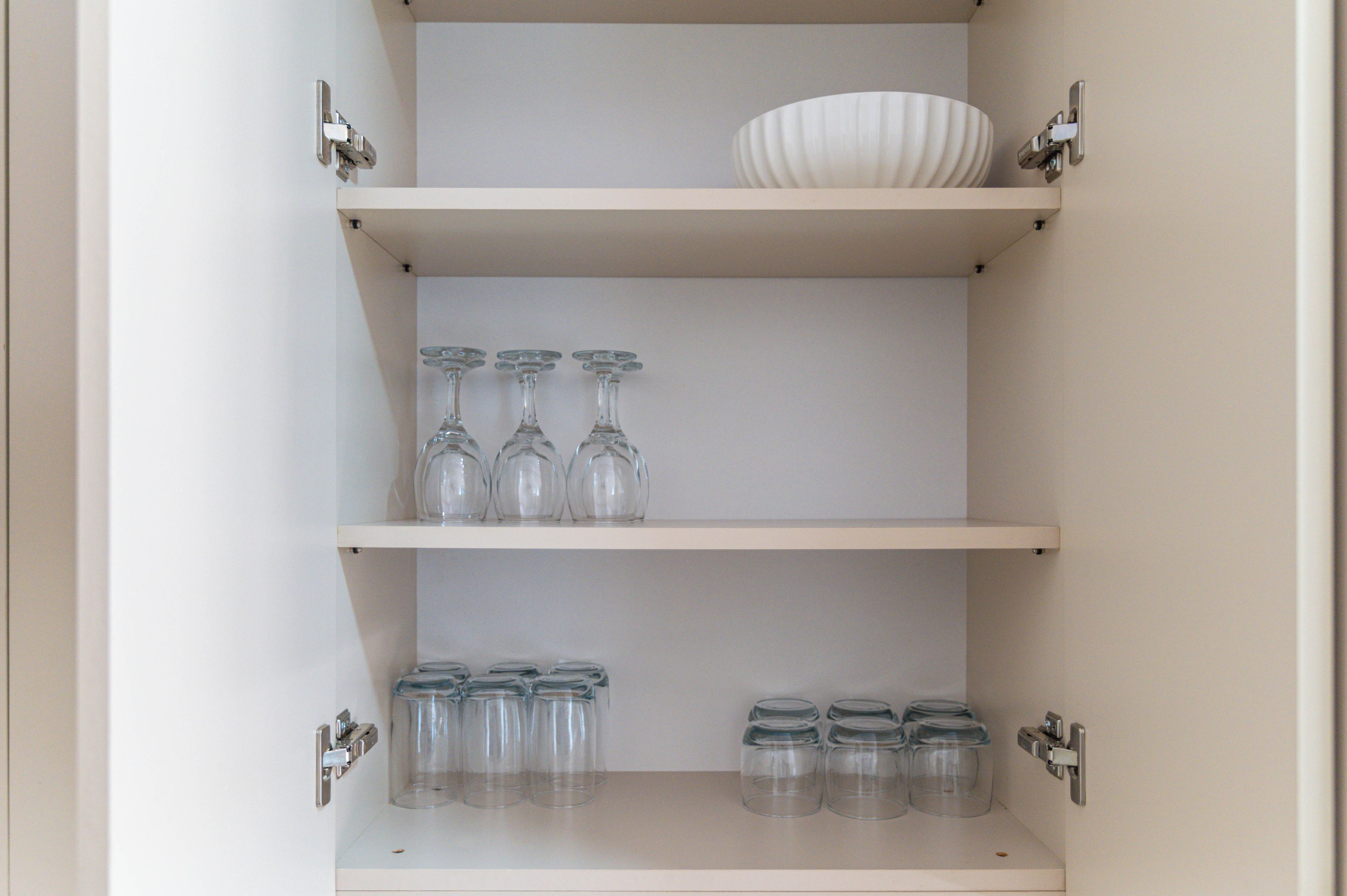 A white shelf unit with a bowl on the top shelf, three wine glasses on the middle shelf, and several clear glass containers on the bottom shelf.