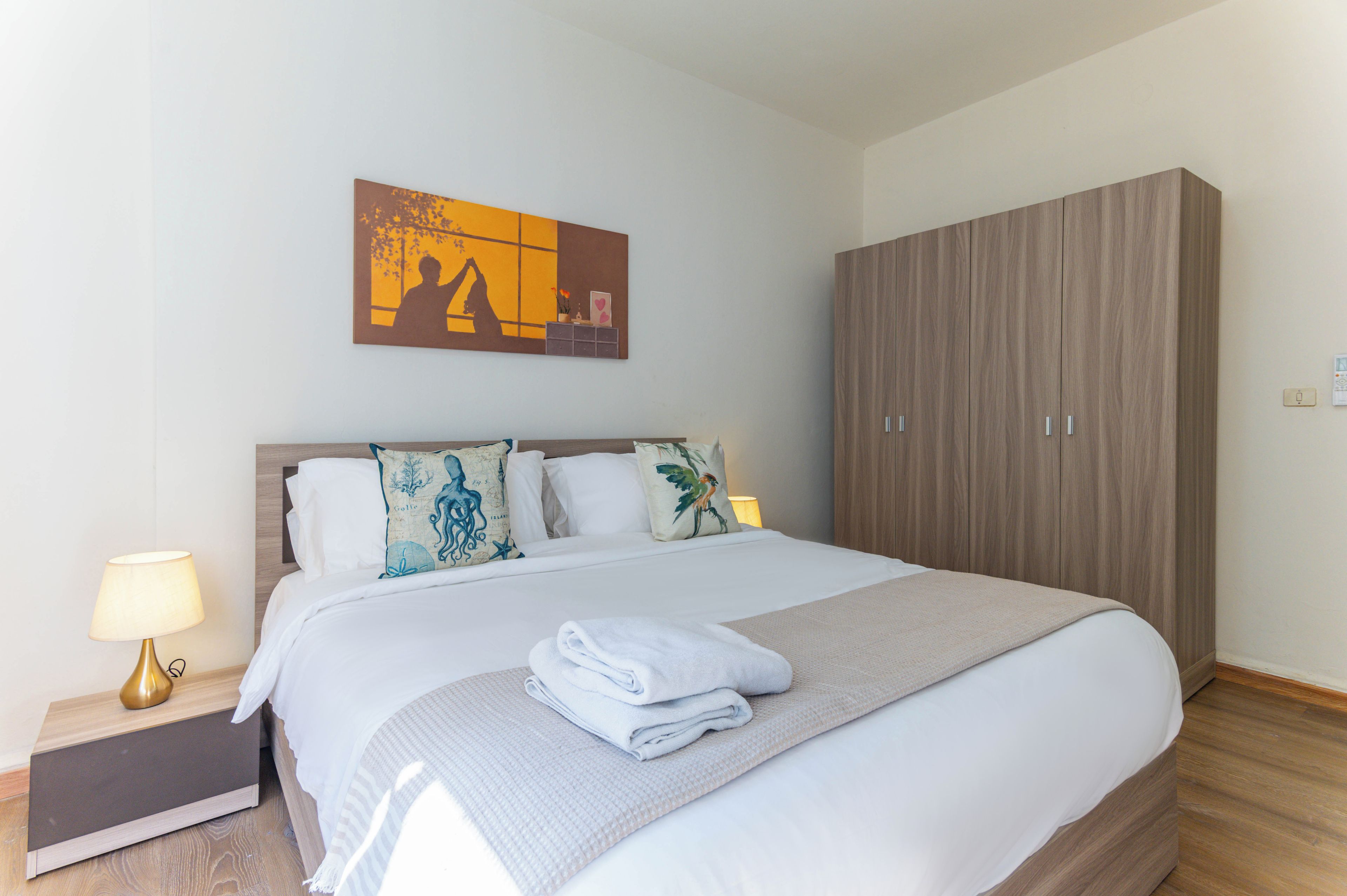 Habitación de hotel con cama, mesita de noche, lámpara, armario y un cuadro en la pared.