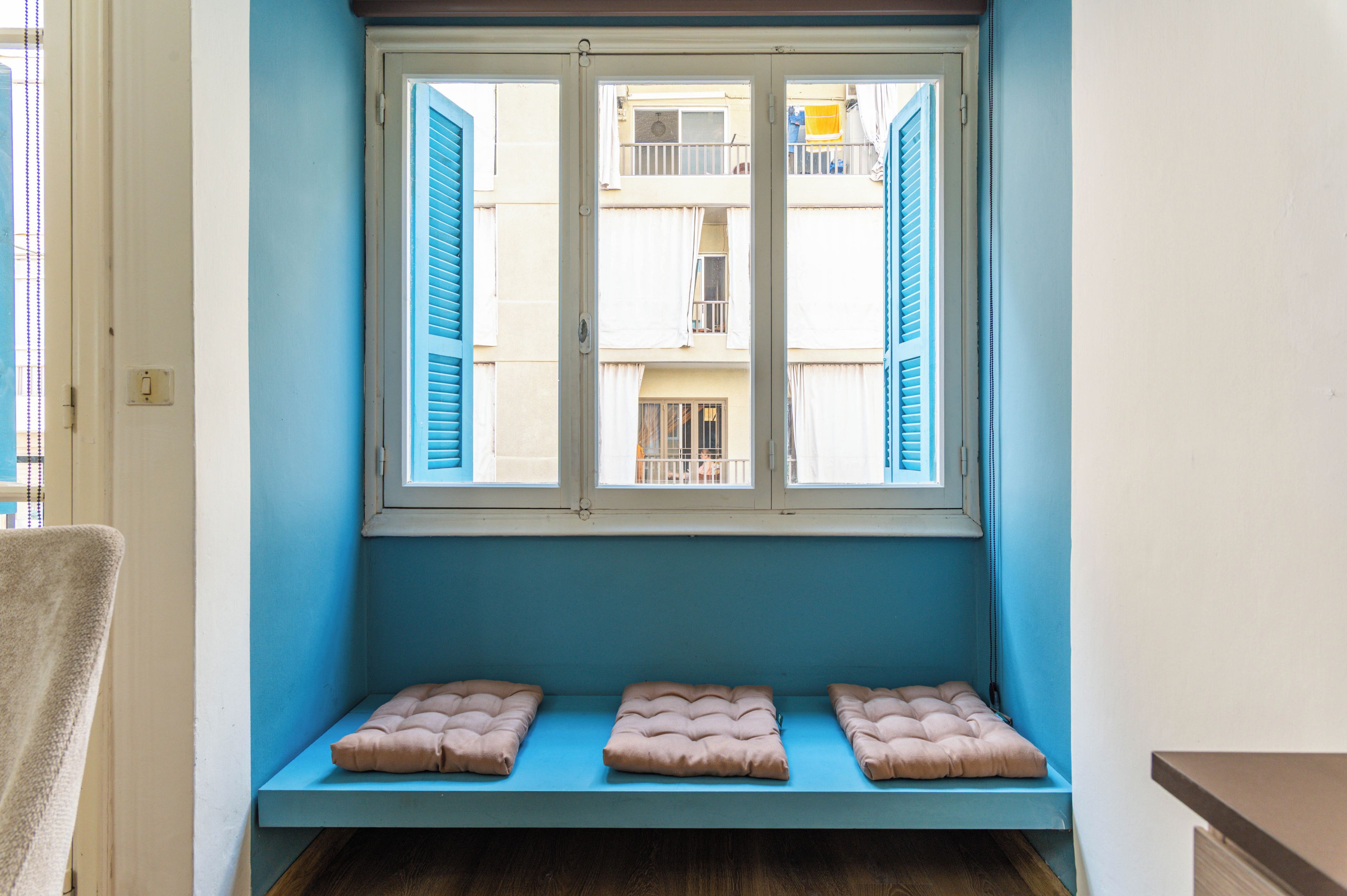 A room with a window overlooking a building with balconies, a bench with cushions, and a towel on the floor.
