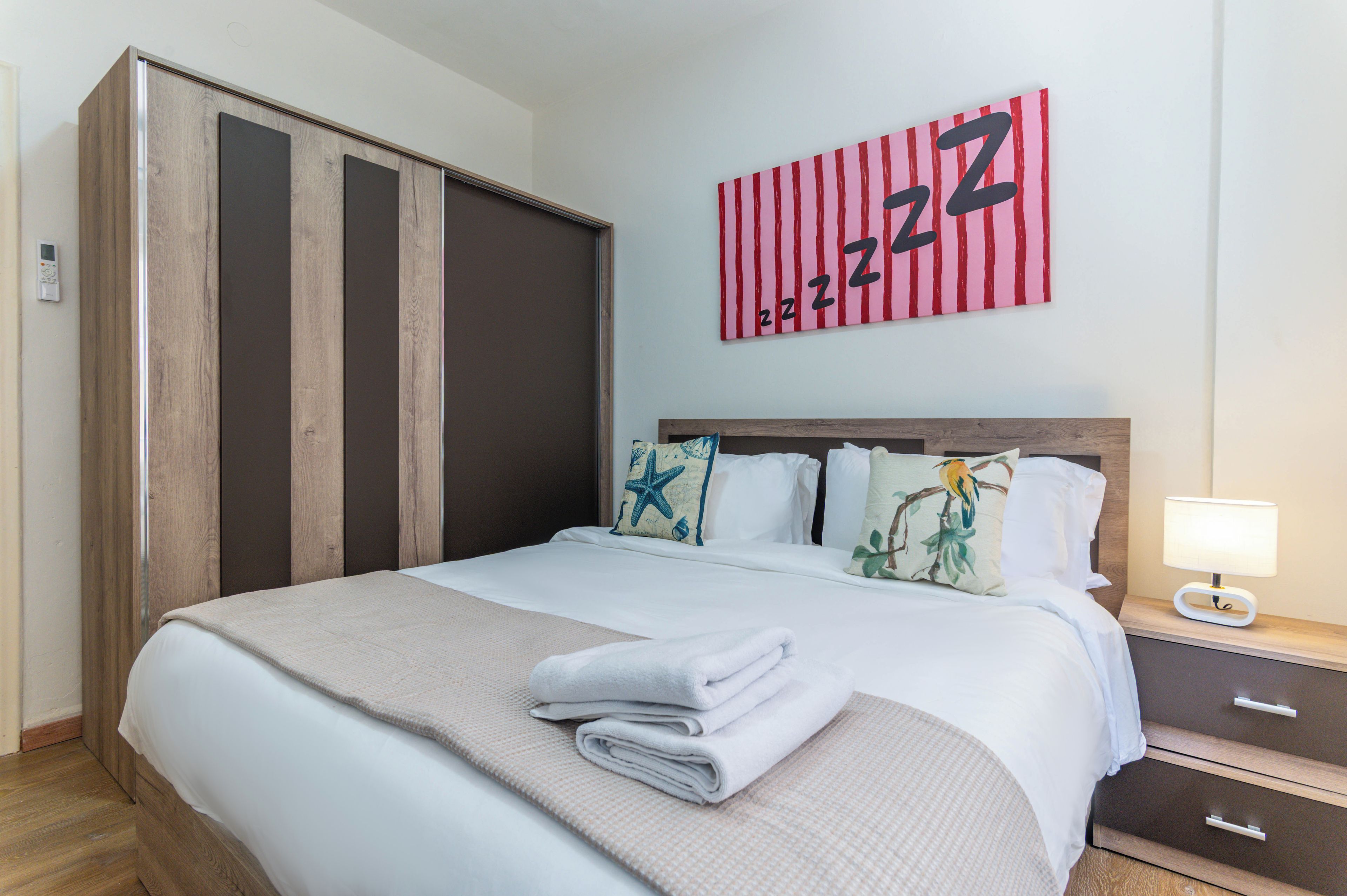 A hotel room with a bed, a nightstand, a lamp, a wardrobe, and a wall with a red and white striped artwork.