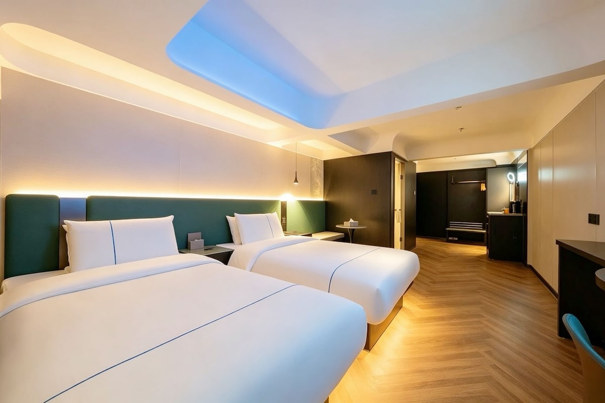 Habitación de hotel con dos camas, tragaluz, escritorio y baño a la vista a través de una puerta abierta.