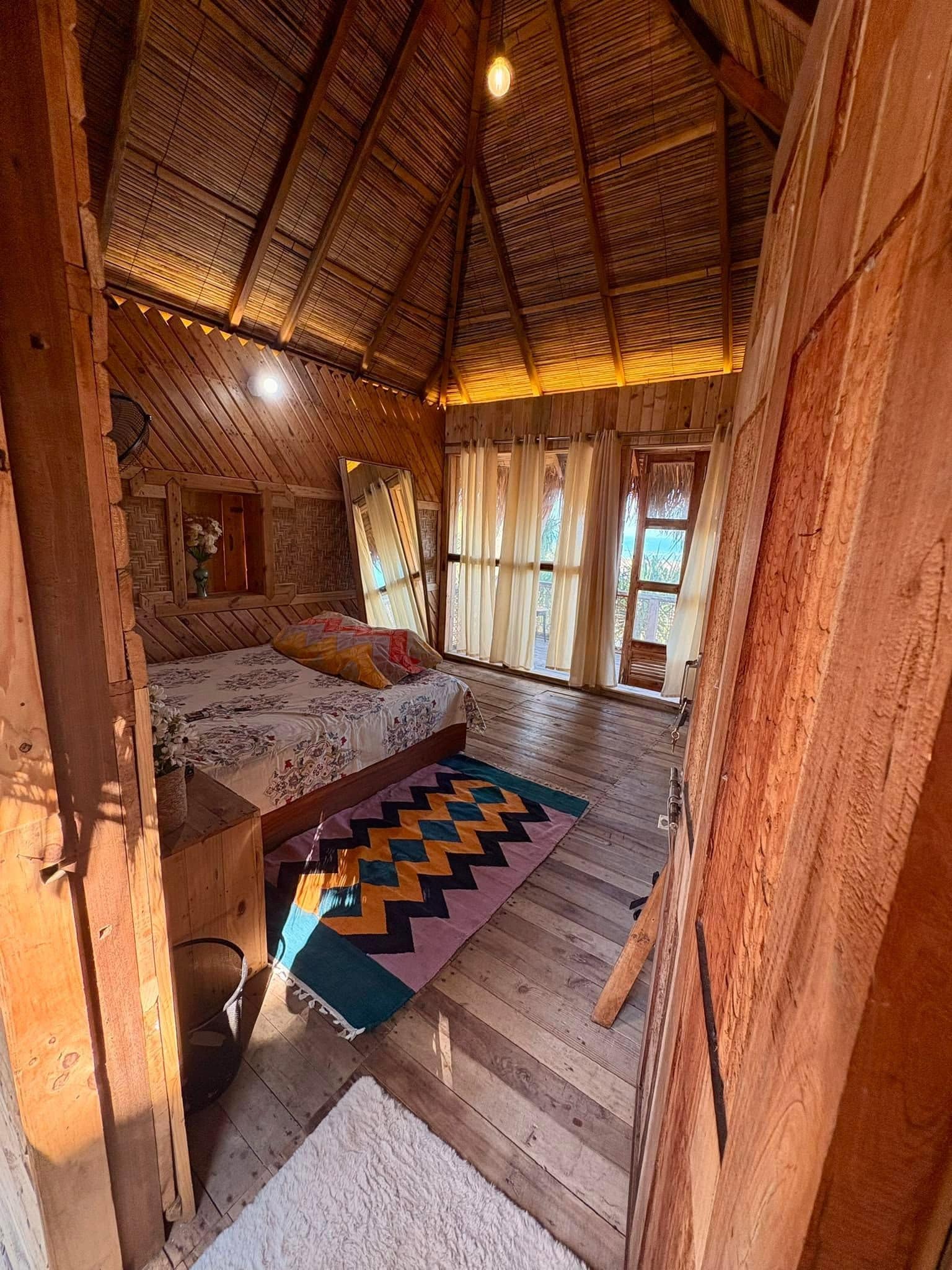 Une pièce rustique en bois, avec un plafond en pente, un lit recouvert d’une courtepointe à motifs et une grande fenêtre ornée de rideaux.