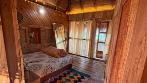 Una habitación rústica de madera con techo inclinado, una cama con colcha estampada y un ventanal con cortinas.