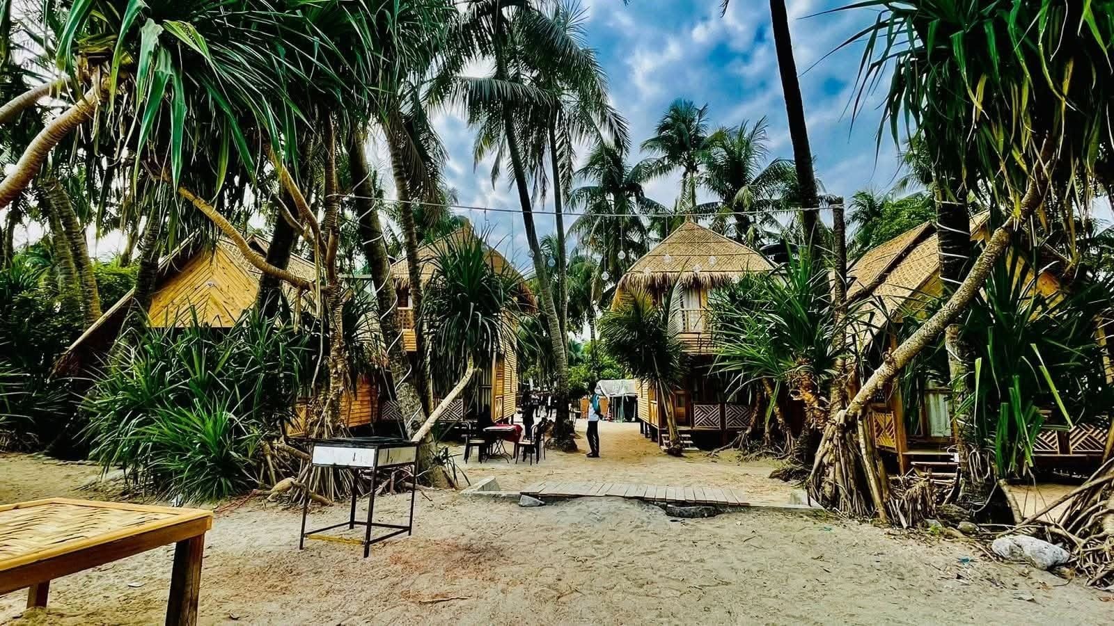 Un complexe hôtelier tropical doté de bungalows aux toits de chaume, de palmiers et d’un sol sablonneux.