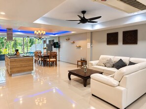 A modern living room with a white sofa, a wooden dining table, and a kitchen area with a stove. - Golf view, pool villa. 5 Bed/Bath. 7 kms to Patong. Peaceful & private (Tambon Kathu)