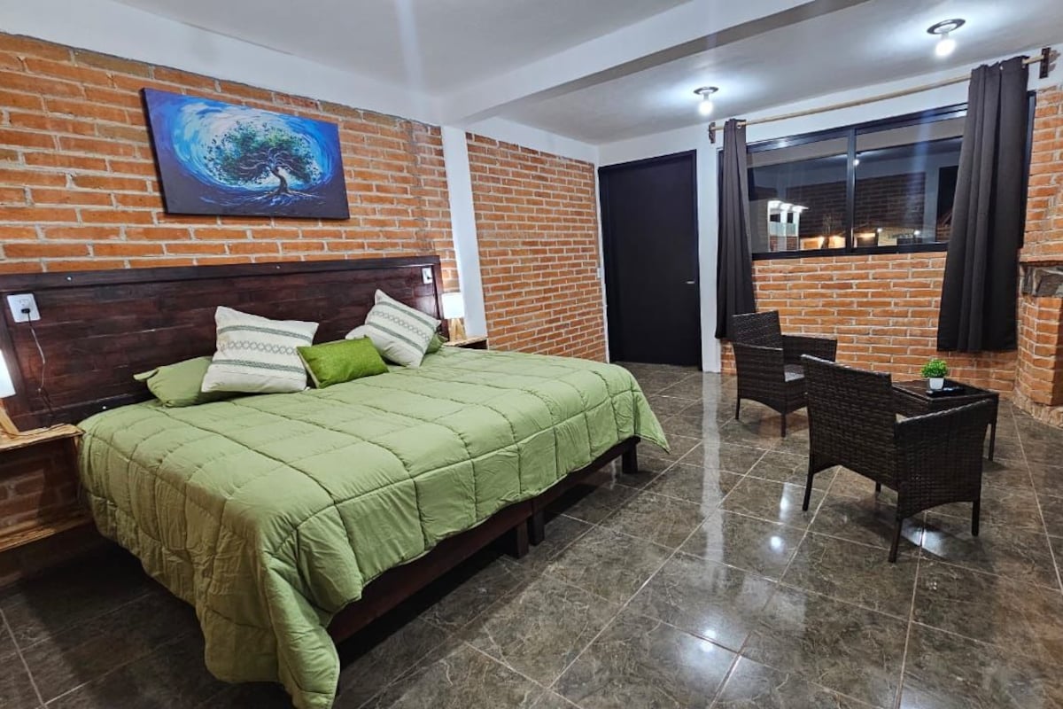 Habitació d'hotel amb paret de maó, llit amb roba de llit verda, una llar de foc, zona de menjador amb taula i cadires i una finestra amb cortines.