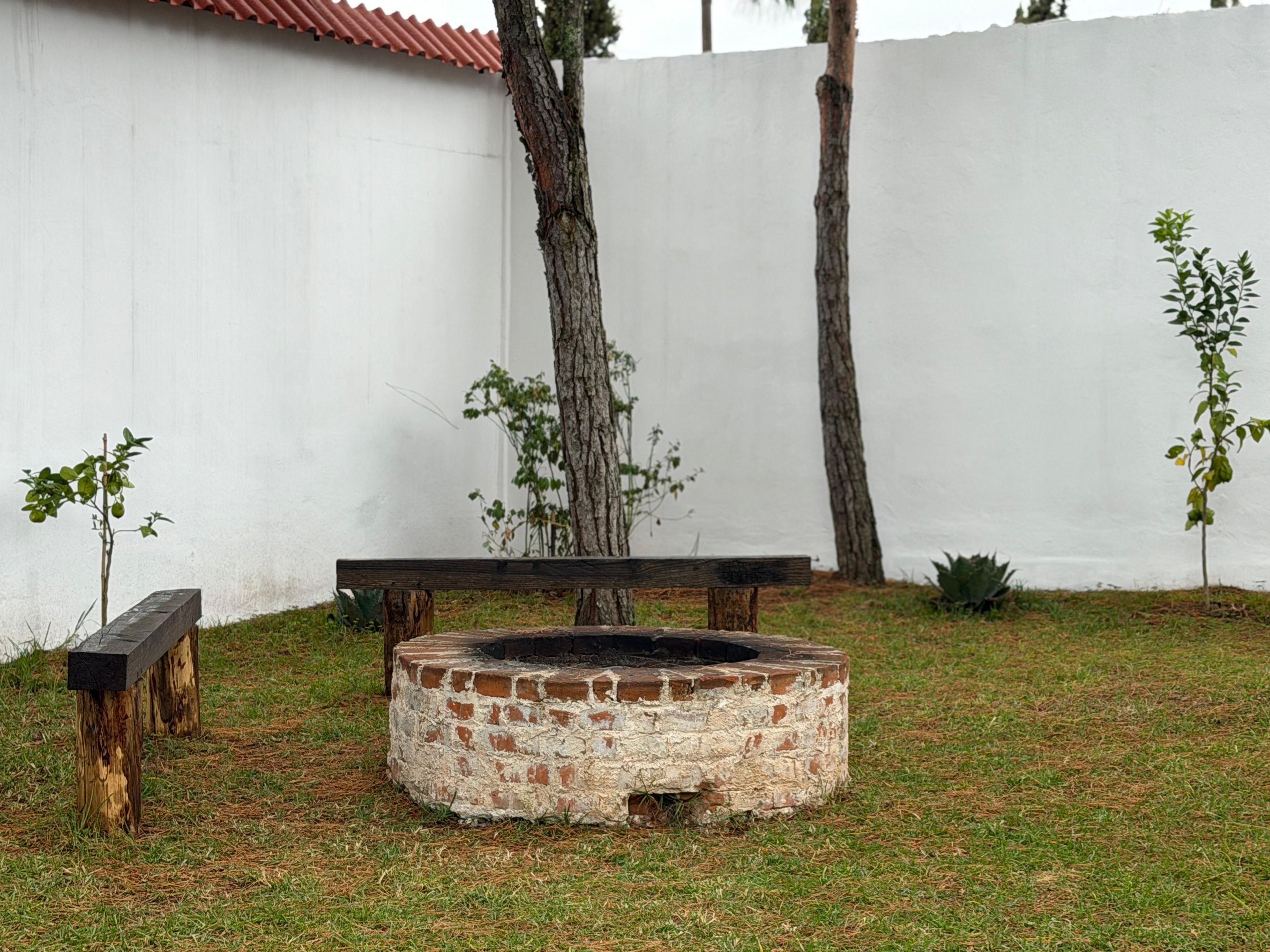 Una chimenea circular con revestimiento de ladrillo, rodeada de césped, un banco de madera y una pared blanca al fondo.