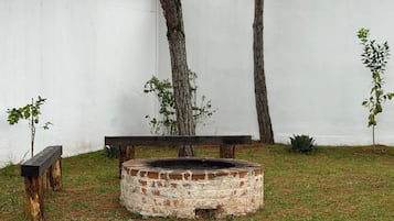 Una chimenea circular con revestimiento de ladrillo, rodeada de césped, un banco de madera y una pared blanca al fondo.