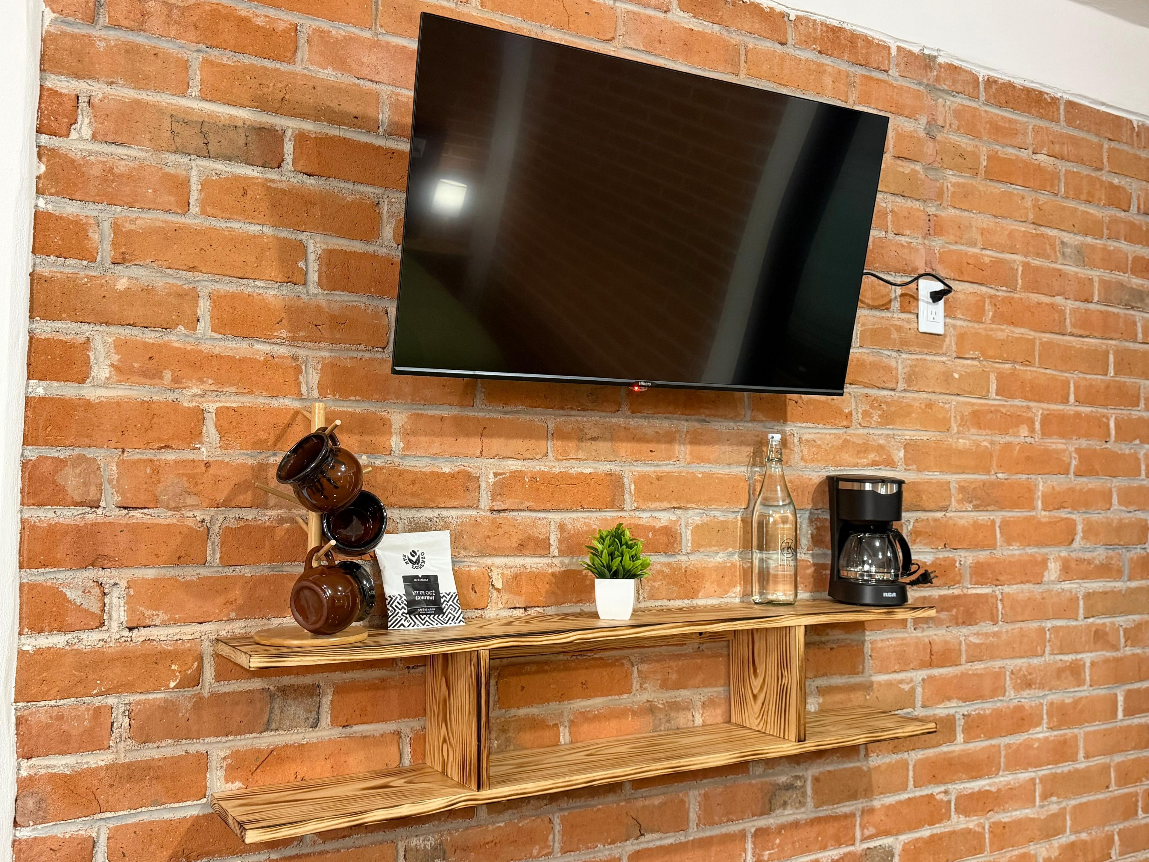 A flat-screen TV mounted on a brick wall, with a wooden shelf below holding a coffee maker, a small potted plant, and decorative items.