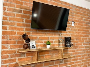 A flat-screen TV mounted on a brick wall, with a wooden shelf below holding a coffee maker, a small potted plant, and decorative items.