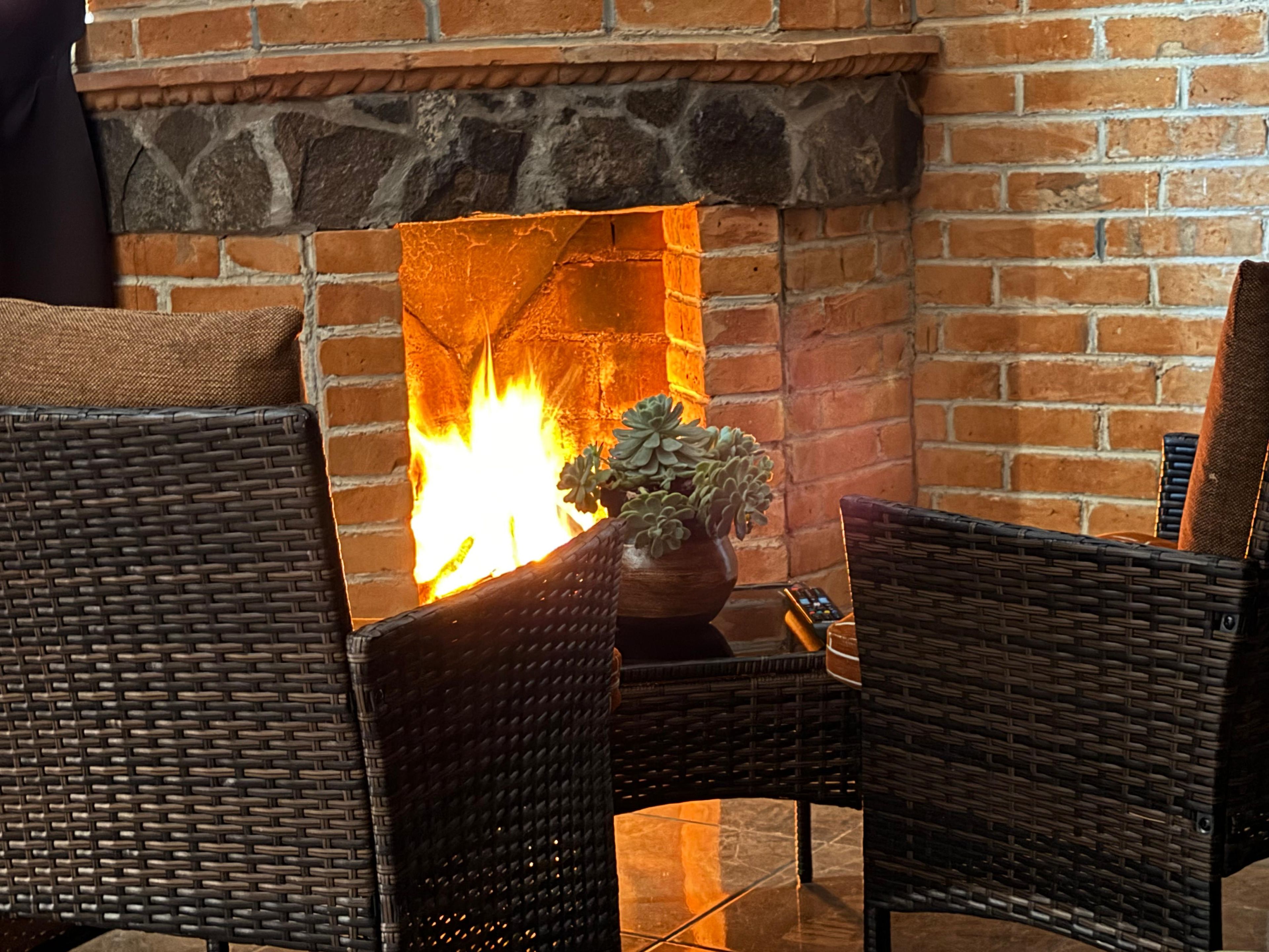 A brick fireplace with a fire, surrounded by wicker furniture.