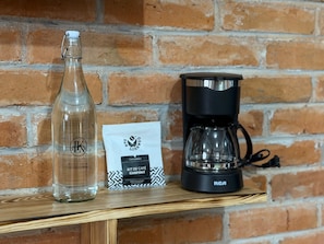 Eine transparente Glasflasche und eine schwarze Kaffeemaschine auf einem Holzboden vor einer Ziegelwand.