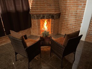 A corner seating area with wicker chairs and a small table, a brick fireplace with a fire, and a curtain on the left. - CABAÑAS LK (Chignahuapan)