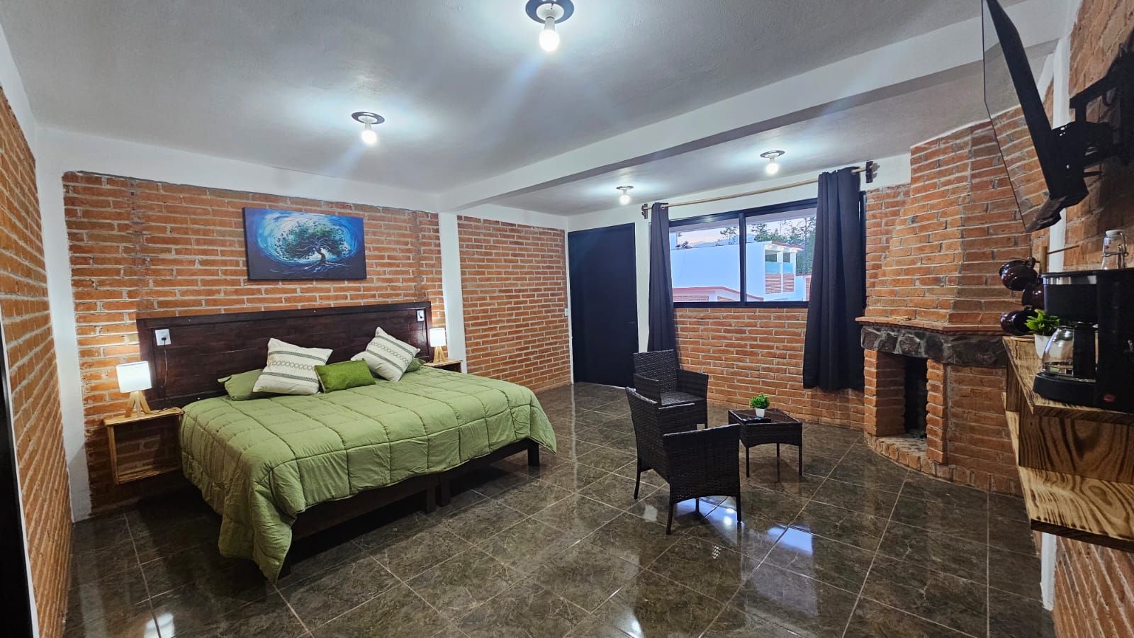 A hotel room with a brick wall, a bed with green bedding, a small table with a plant, and a fireplace.