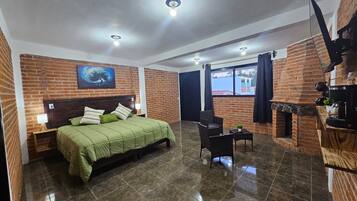 A hotel room with a brick wall, a bed with green bedding, a small table with a plant, and a fireplace.