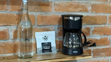 A clear bottle, a coffee maker, and a gift bag on a wooden shelf against a brick wall.