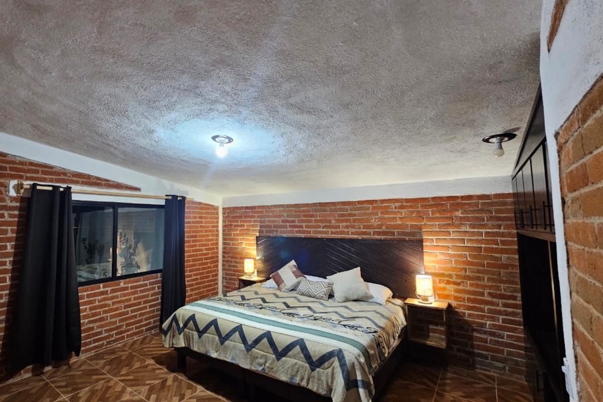 Un dormitorio con pared de ladrillo, cama con colcha estampada, mesita de noche con lámpara y ventana con cortinas.