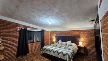 A bedroom with a brick wall, a bed with a patterned bedspread, a nightstand with a lamp, and a window with curtains.