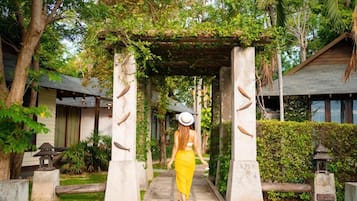 A pathway leading through a garden with a covered walkway, surrounded by greenery and a building with a sloped roof.