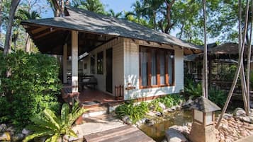 Una casa de madera con porche, rodeada de follaje tropical y un pequeño estanque.
