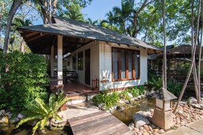 Een houten huis met veranda, omringd door tropische begroeiing en een klein vijvertje.