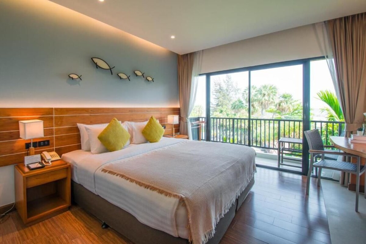 Een hotelkamer met een groot bed, een nachtkastje met telefoon, een balkon met tafel en stoelen, en uitzicht op groen.
