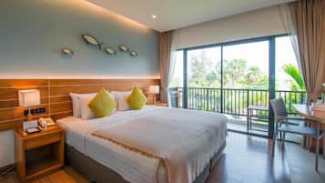 A hotel room with a large bed, a nightstand with a phone, a balcony with a table and chairs, and a view of greenery outside.