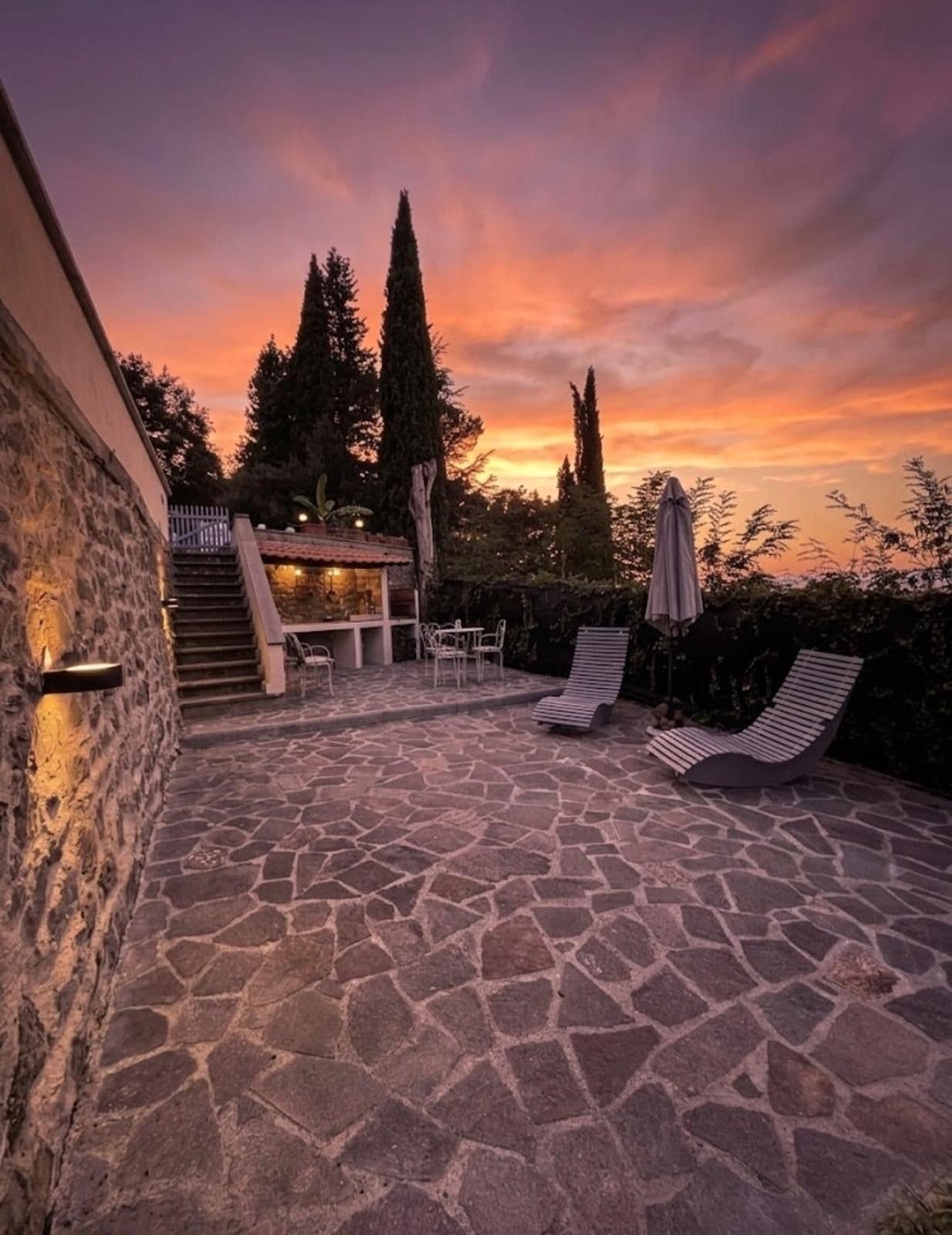 Une terrasse en pierre avec des chaises longues et une table, entourée d’arbres et d’un bâtiment doté d’un escalier.