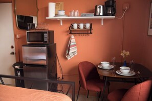 A compact kitchen with a microwave, toaster, and coffee maker on a shelf, a dining table with chairs, and a small table with a vase of flowers.