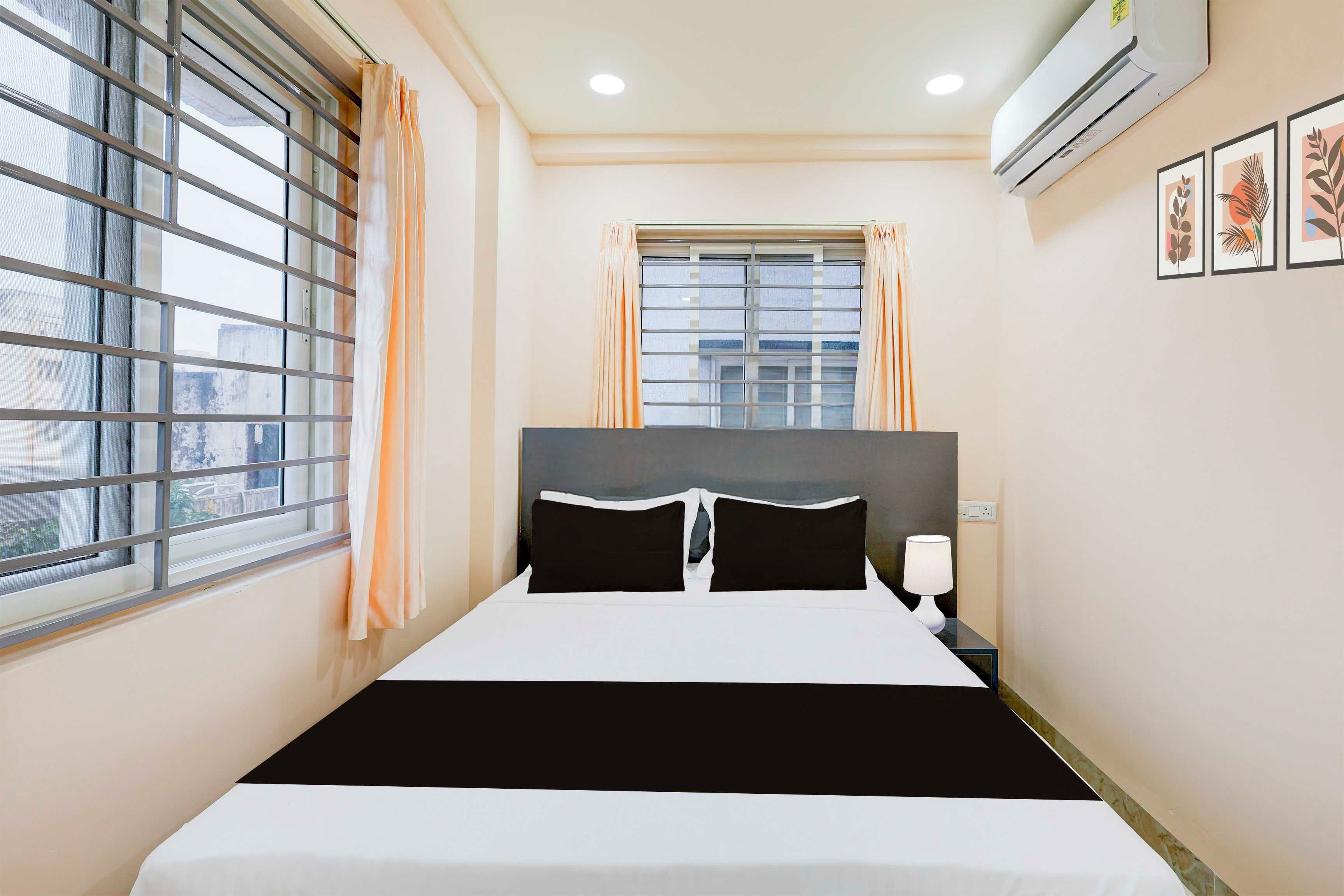 A hotel room with a bed, a window with curtains, an air conditioner, and a wall-mounted lamp.
