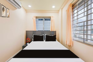 A bedroom with a bed, a window with blinds, a nightstand, and a vase with flowers. - Townhouse Oak Visakhapatnam Lumbini Park (Visakhapatnam)